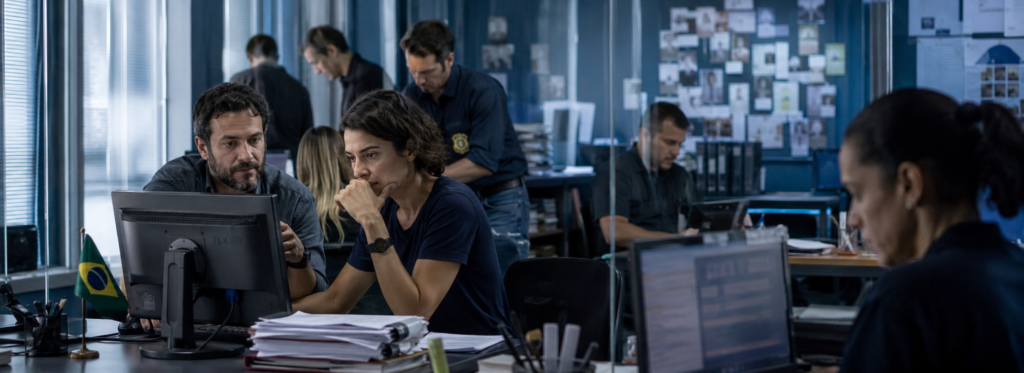 Banner horizontal com fotografia corporativa realista em tons de azul mostrando uma equipe de investigadores e analistas reunida em um escritório moderno no Brasil. Em primeiro plano, um homem e uma mulher analisam informações em um computador com expressão de concentração, enquanto ao fundo outros profissionais trabalham entre documentos, monitores e um painel de investigação. A imagem remete ao universo de filmes sobre corrupção no Brasil, com atmosfera séria, investigativa e institucional.