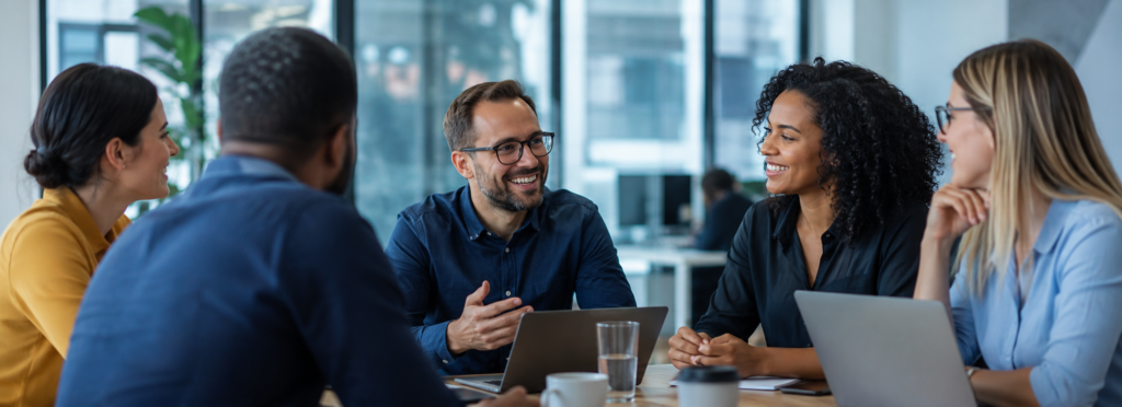 Equipe diversa em reunião em um escritório moderno, conversando de forma colaborativa ao redor de uma mesa com notebooks, em ambiente corporativo iluminado por luz natural.
