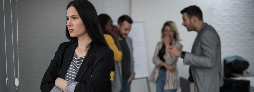 mulher branca em primeiro plano e colegas ao fundo conversando sobre ela