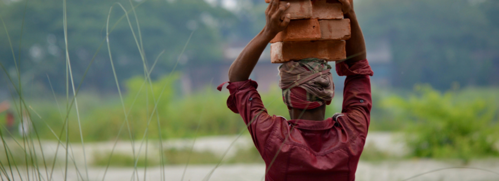 Trabalhador com lenço na cabeça carrega uma pilha de tijolos sobre a cabeça, visto de costas, em área externa com vegetação ao fundo.