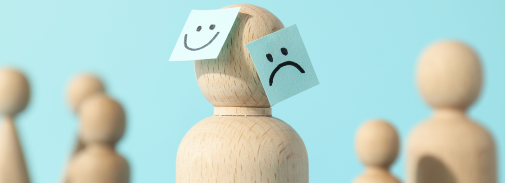 Boneco de madeira com rostos feliz e triste representando riscos psicossociais e saúde mental no trabalho, tema da NR-01